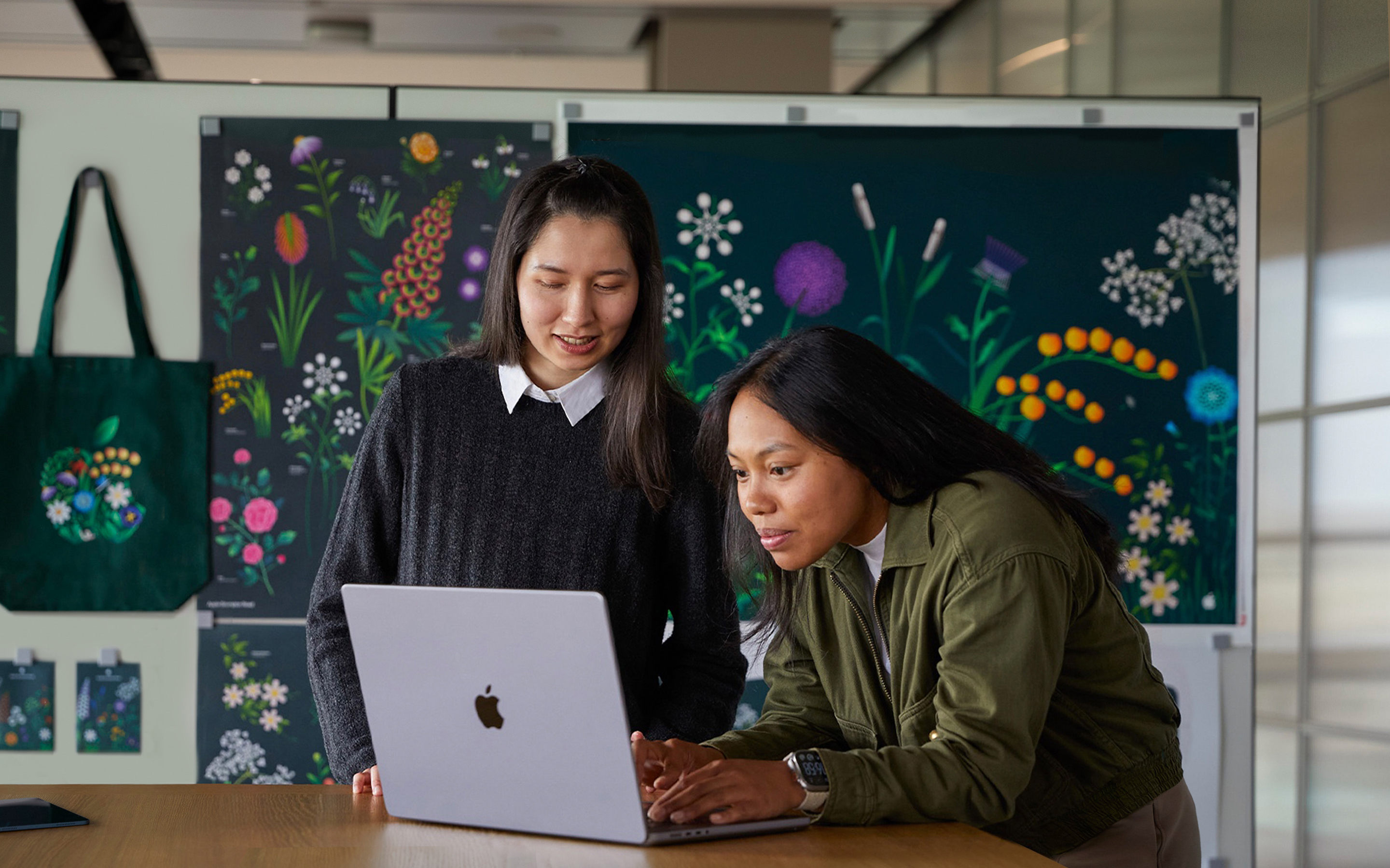 To ansatte på Apples markedsføringsteam samarbeider på en MacBook foran plakater designet av teamet.