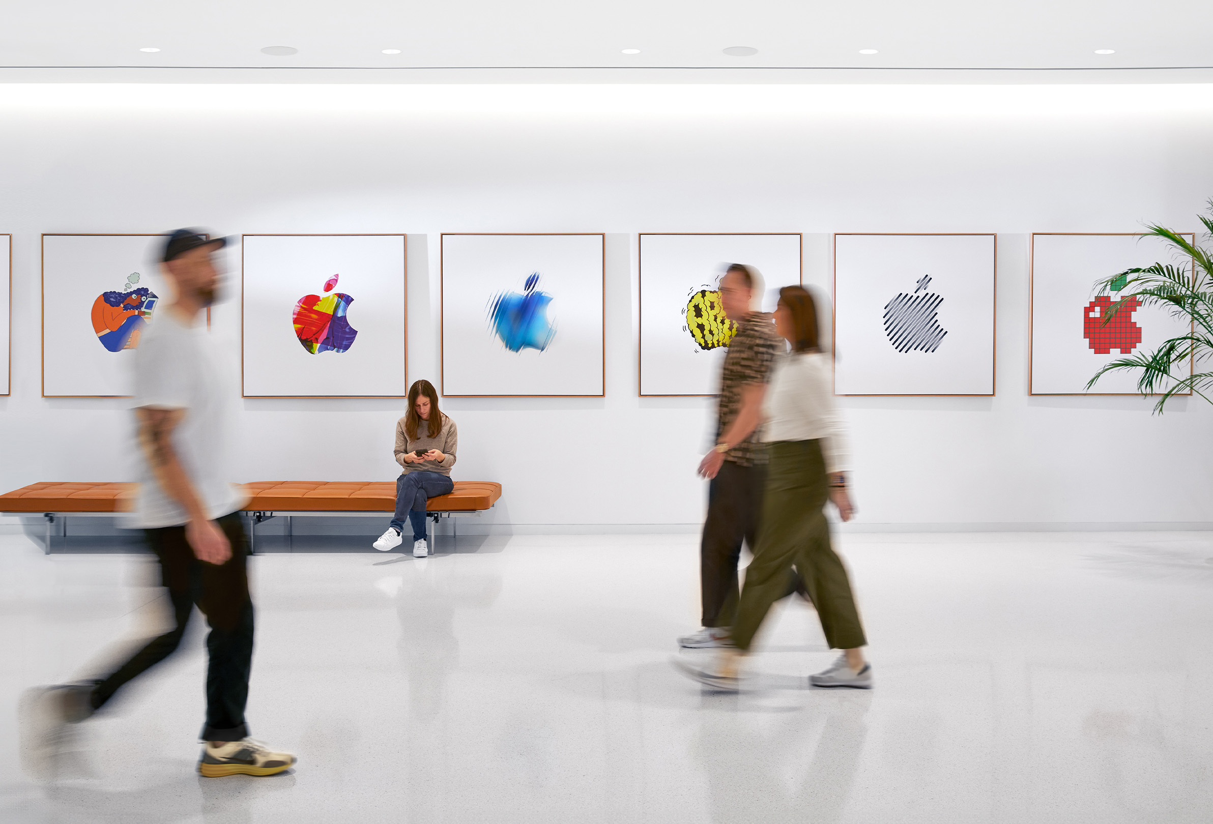 Three Apple colleagues walking past an interior wall with colourful Apple logos and a fourth employee sitting on a bench.
