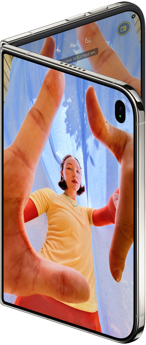 Pootevřený nebesky bílý iPhone&nbsp;Duo, na vnějším displeji je aplikace Fotoaparát a&nbsp;Center Stage přední kamera, na vnitřním knihovna aplikace Fotky