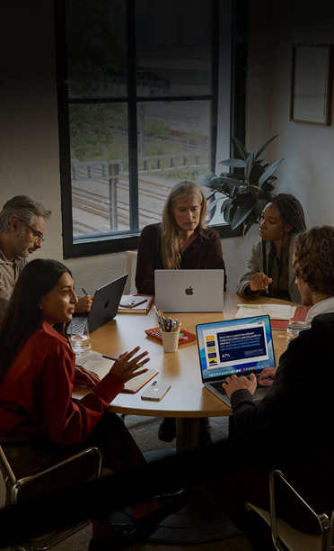 Five individuals seated around a conference table, collaborating on MacBook computers, with iPhones face down on the table