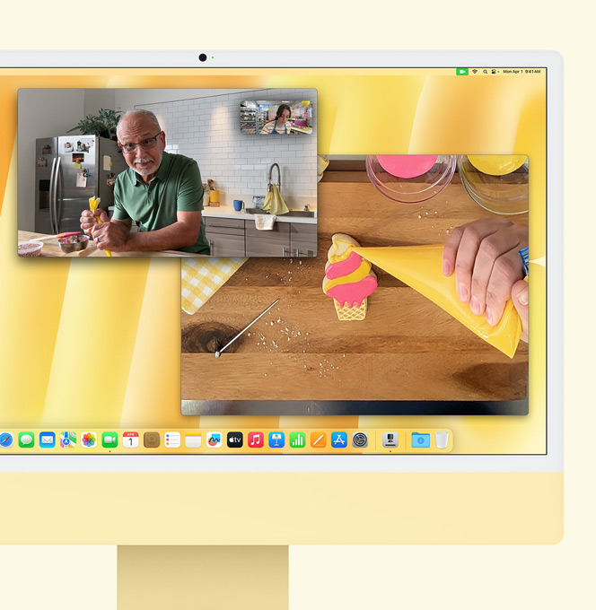 Yellow iMac. Desk View demo. Screen shows baker on video call with another person. One screen displays the baker and the man’s face. The other screen displays the baker’s hands icing a biscuit