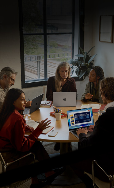 Five individuals seated around a conference table, collaborating on MacBooks, with iPhones face down on the table