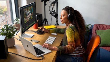 امرأة في منزلها تعمل خلف مكتبها مع MacBook Pro متصل بشاشتين خارجيتين وترتدي سماعات رأس وتتحدث في الميكروفون