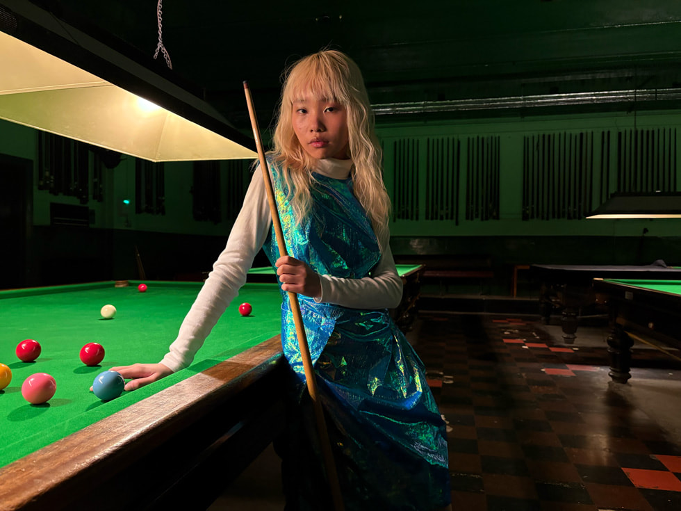 Loş ışıklı bir odada bilardo masasının karşısında poz veren bir kişi.