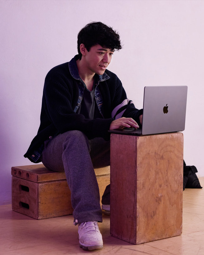 Kyle Hanagami sentado sobre una caja en un estudio escribe en una MacBook Pro ubicada sobre otra caja.
