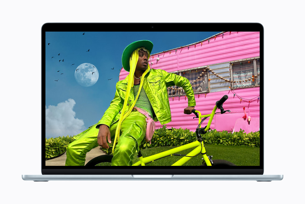 Una MacBook Air muestra la foto de una persona con un atuendo verde brillante sentada en una bicicleta del mismo color frente a una casa rodante rosa brillante.