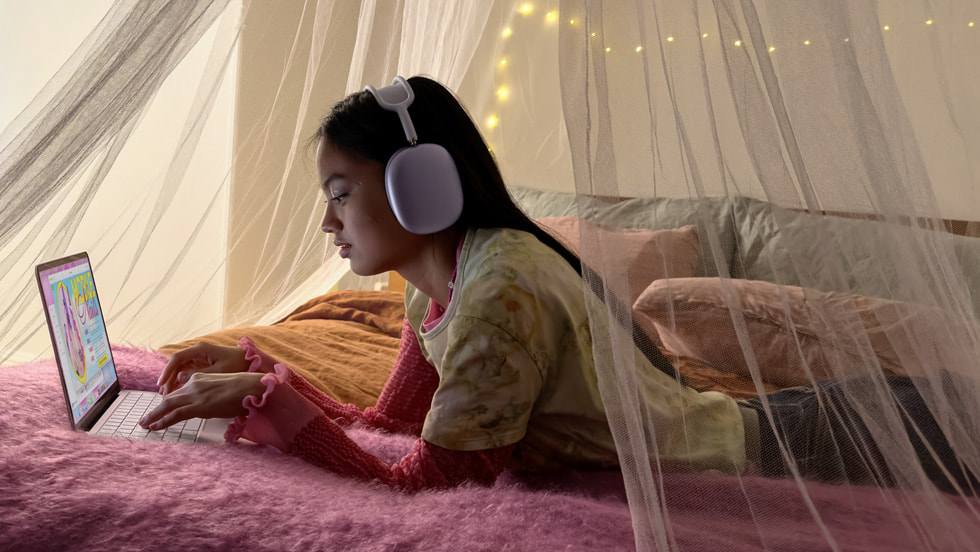 Una persona recostada en su cama usa una MacBook Neo mientras escucha música con unos AirPods Max.