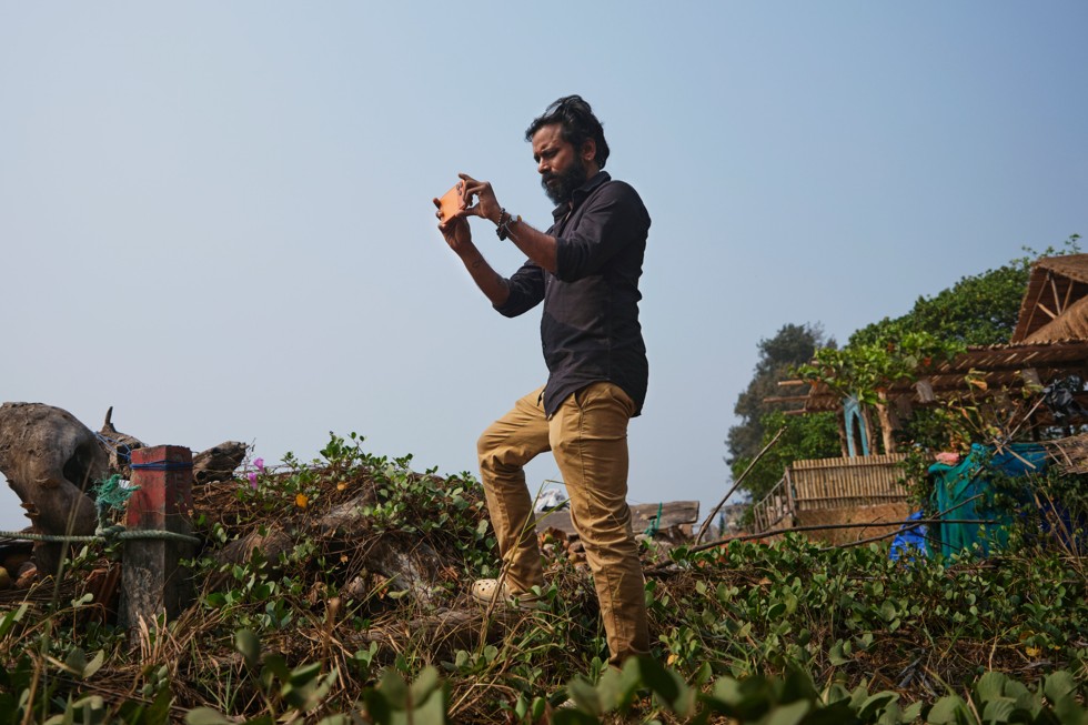 Ritesh Sharma de pie en un montículo cubierto de hojas sostiene un iPhone 17 Pro Max mientras graba mirando hacia el mar.