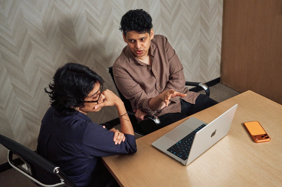 Shreela Agarwal sentada en una mesa hablando con el mentor Chaitanya Tamhane, con una MacBook Pro y un iPhone 17 Pro Max apoyado sobre la mesa.