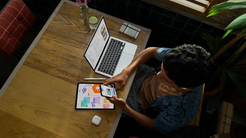 Vista superior de una persona que sostiene un iPhone y trabaja en una mesa con una MacBook Pro en la que se muestra la programación en la pantalla, junto con un iPad, un Apple Pencil y unos AirPods.