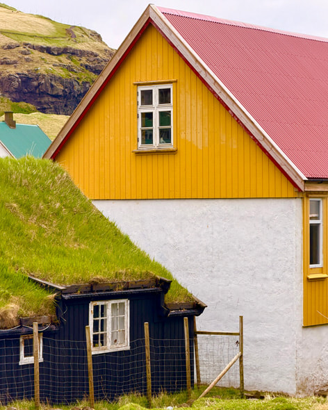 A jewel-toned yellow and pink building sits behind a dark house with a traditional grass roof in the Faroe Islands. Shot on iPhone 17 Pro by Anastasiia Sapon.