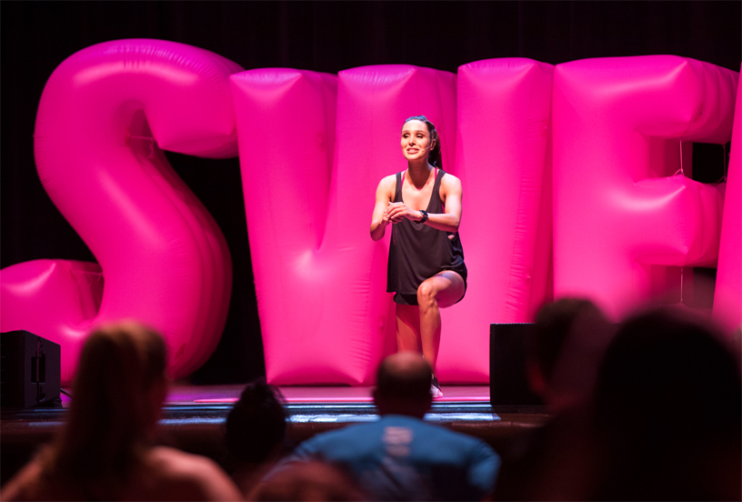 Kayla Itsines sahnede şişirilmiş balon harfler önünde fitnes rutinini yapıyor. 