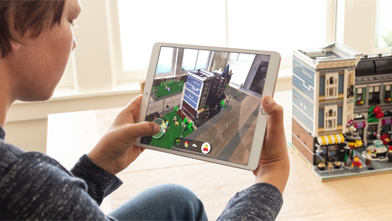 Boy sitting in front of a LEGO building in a room, holding an iPad with LEGO AR City on the screen. 