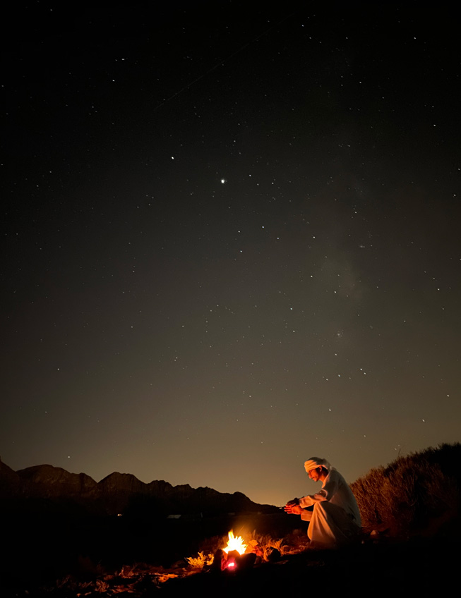 Gece çölde kamp ateşinin başında oturan bir adamın fotoğrafı.