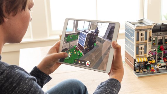 Ekranında LEGO AR City olan iPad’ini elinde tutarak bir odada LEGO binasının önünde oturan çocuk.