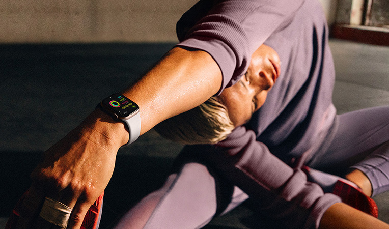 Yerde oturmuş, kolunu bacağına doğru esneten bir kadının bileğindeki Apple Watch Series 10’un ekranında Antrenman, Soğuma, Aktivite ve Aktivite Halkaları görülüyor
