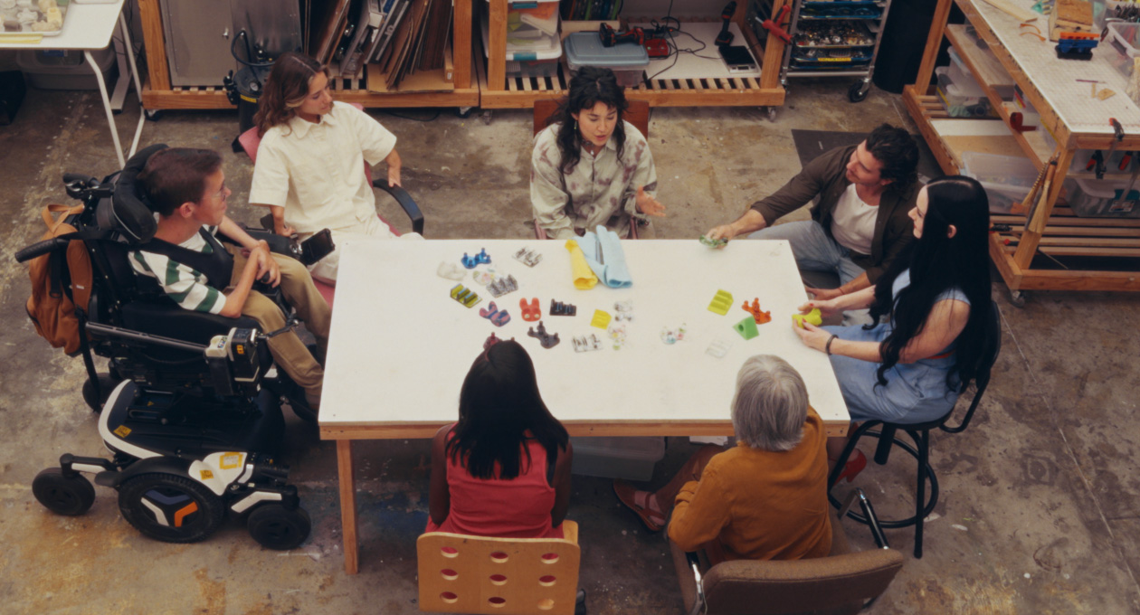 Un grupo de personas con diferentes necesidades de movilidad sentadas en torno a una mesa miran una serie de accesorios adaptados de varios colores para smartphones