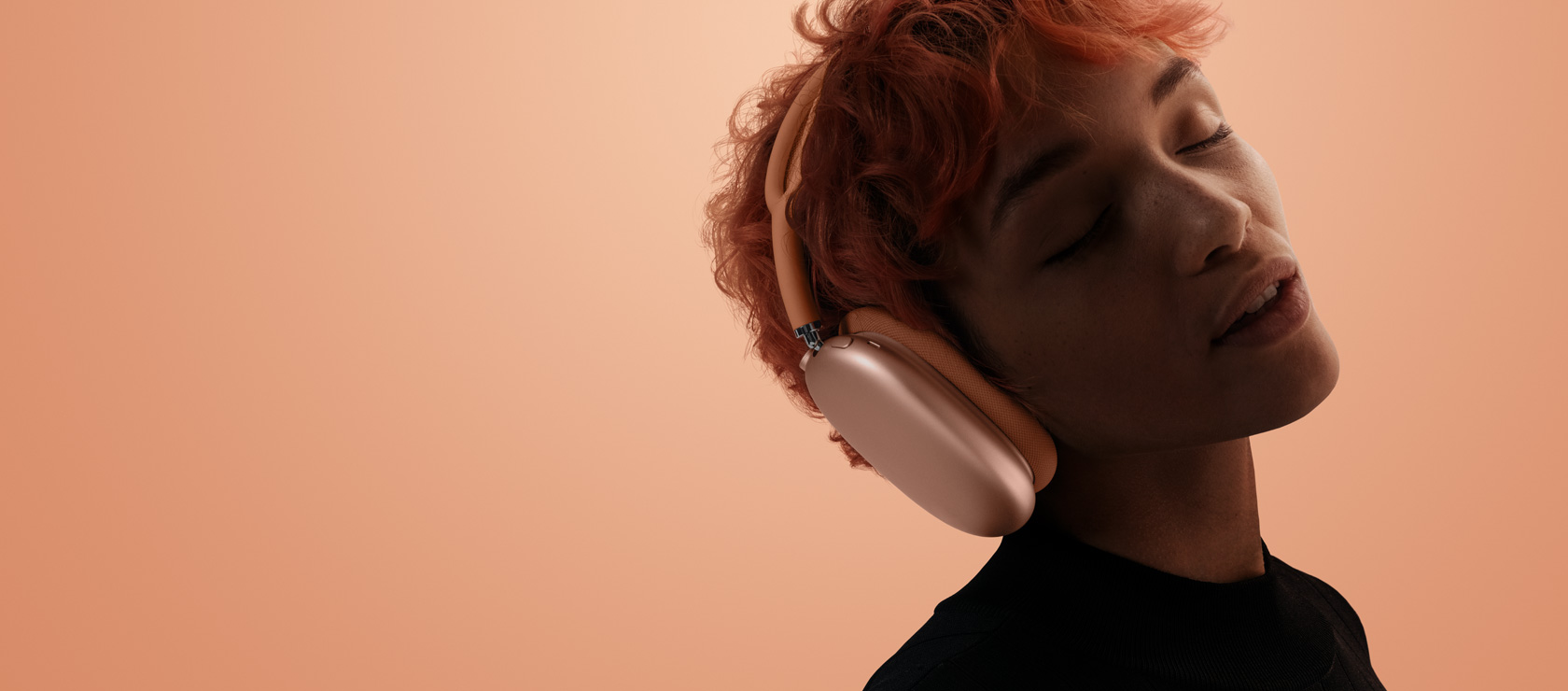 Eine Person mit geneigtem Kopf und geschlossenen Augen, die AirPods Max 2 in Orange trägt, vor einem orange-rosa Hintergrund