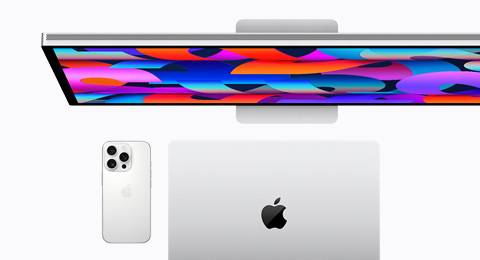 An Apple Display behind an iPhone 16 Pro in White Titanium color and a closed MacBook in Silver color