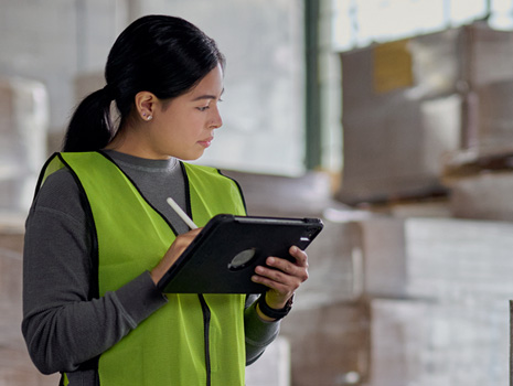 Una mujer de pie en una fábrica con un iPad en la mano mientras controla el inventario. Detrás de ella se muestran pilas y pilas de cajas grandes.