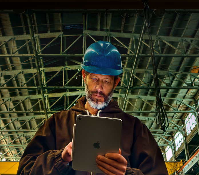iPad, silver, with Apple Pencil attached, held by a person in a brown jacket and blue hard hat, working in an industrial setting with metal beams overhead