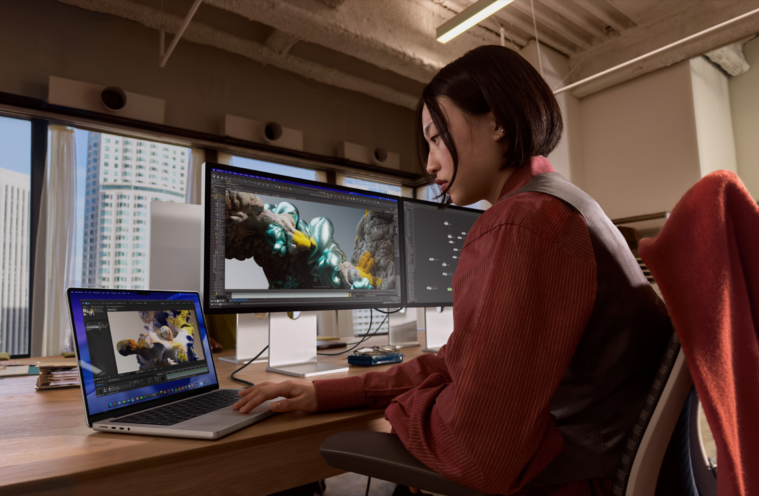 A person works at a desk with a MacBook Pro connected to an external display, both screens showing a 3D render, buildings visible through the window behind