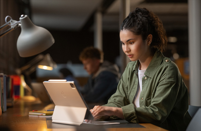 Une étudiante assise à un bureau dans une salle d’étude tape sur un iPad avec un Magic Keyboard, illustrant la polyvalence des appareils Apple