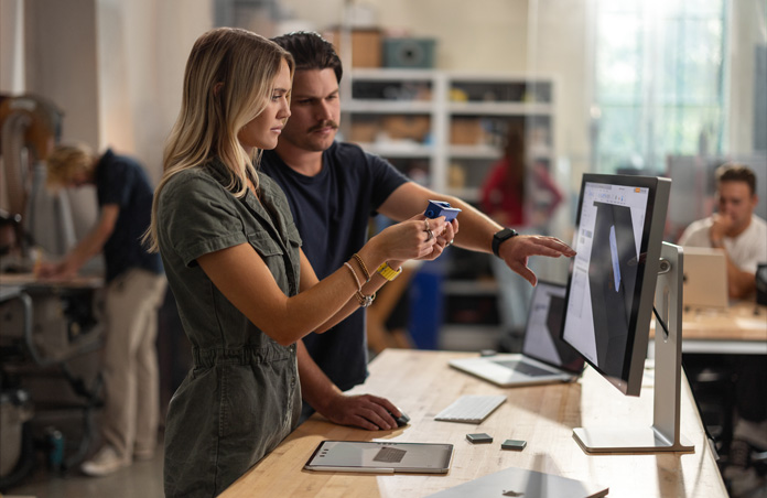 Two students collaborate with a real component model while viewing a 3-D project on a Mac Studio Display, MacBook, Apple Pencil and iPad rest on table.