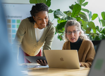 Deux professeures collaborent autour d’un bureau, l’une d’elles tient un iPad tandis que l’autre travaille sur un Mac
