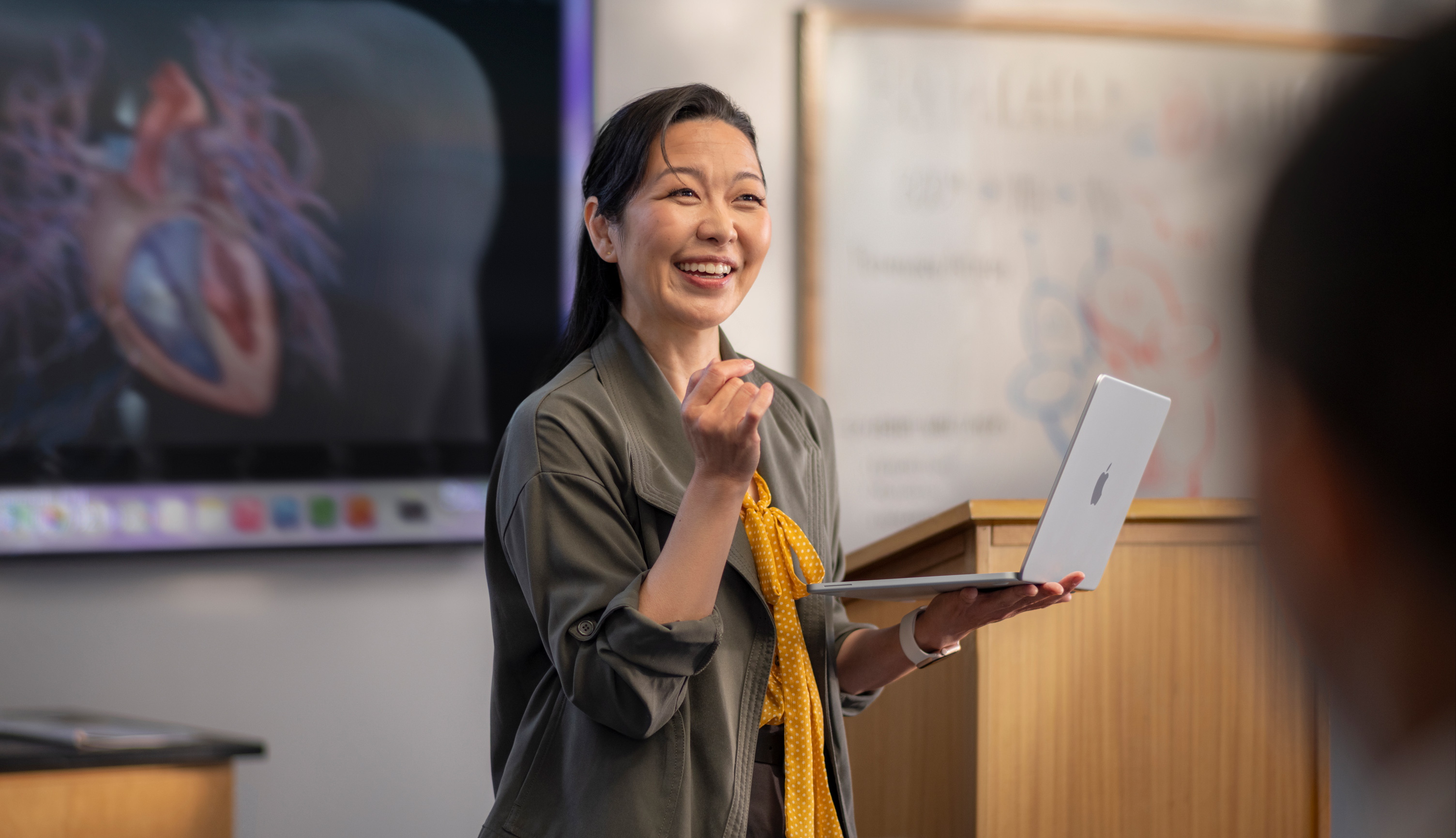 Une professeure donne un cours dans un amphithéâtre tout en tenant un MacBook, illustrant les performances et la grande autonomie de l’appareil