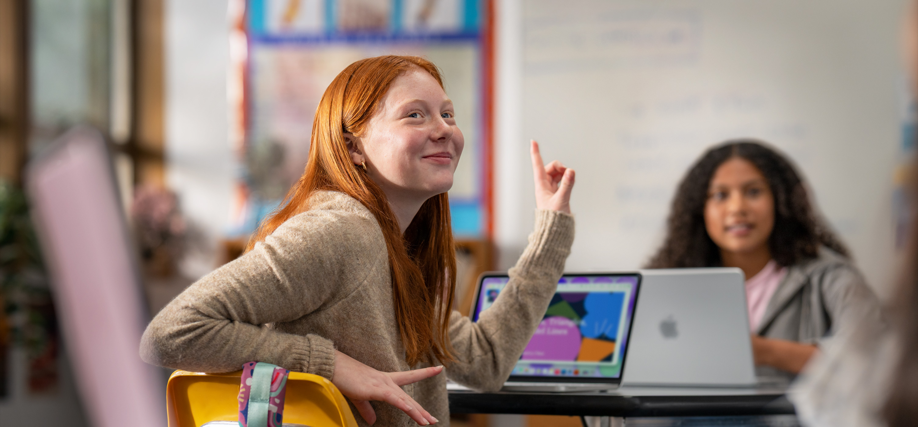 Eine Schülerin sitzt in einem Klassenzimmer vor einem MacBook und hebt die Hand, um eine Frage zu beantworten, was zeigt, dass es einfach zu bedienen und motivierend ist