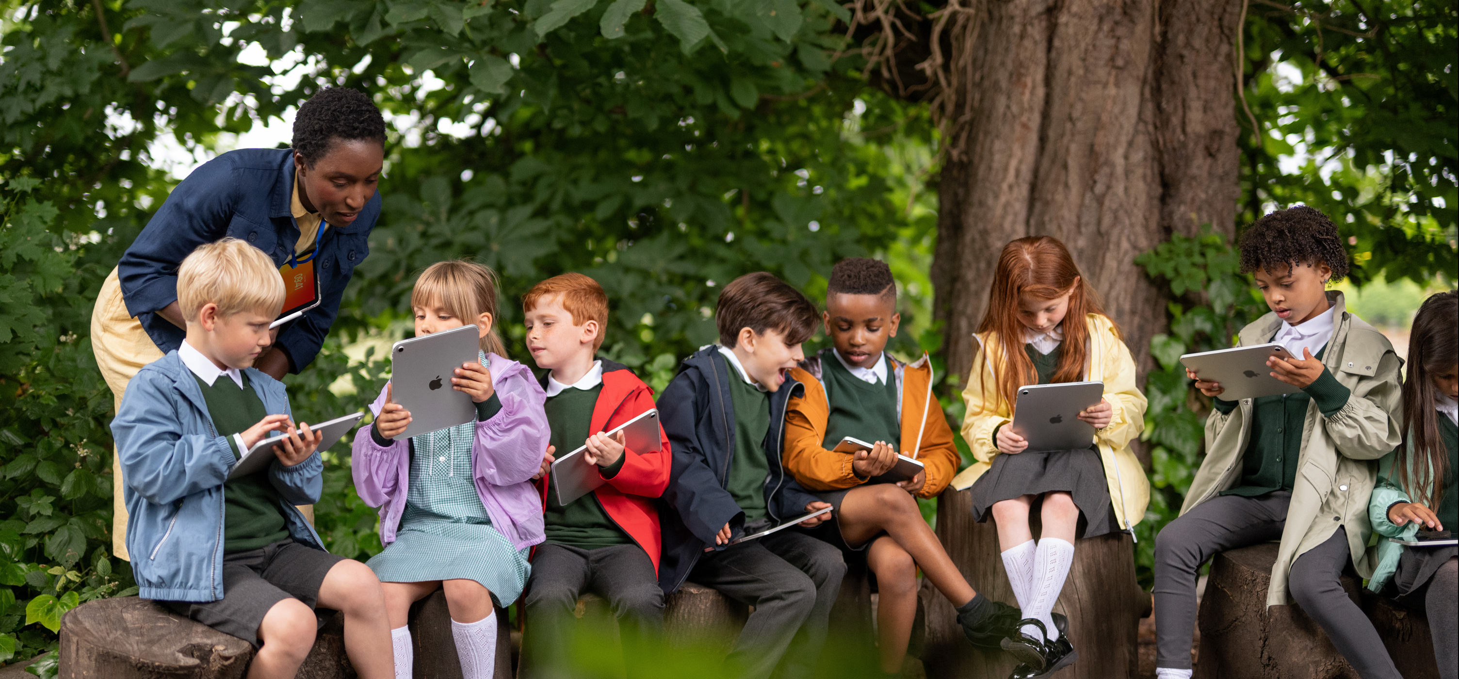 Eine Gruppe junger Schüler:innen nutzt das iPad im Freien, während eine Lehrkraft sie mit einem iPad in der Hand anleitet und so hervorhebt, dass Mobilität das Lernen überall ermöglicht