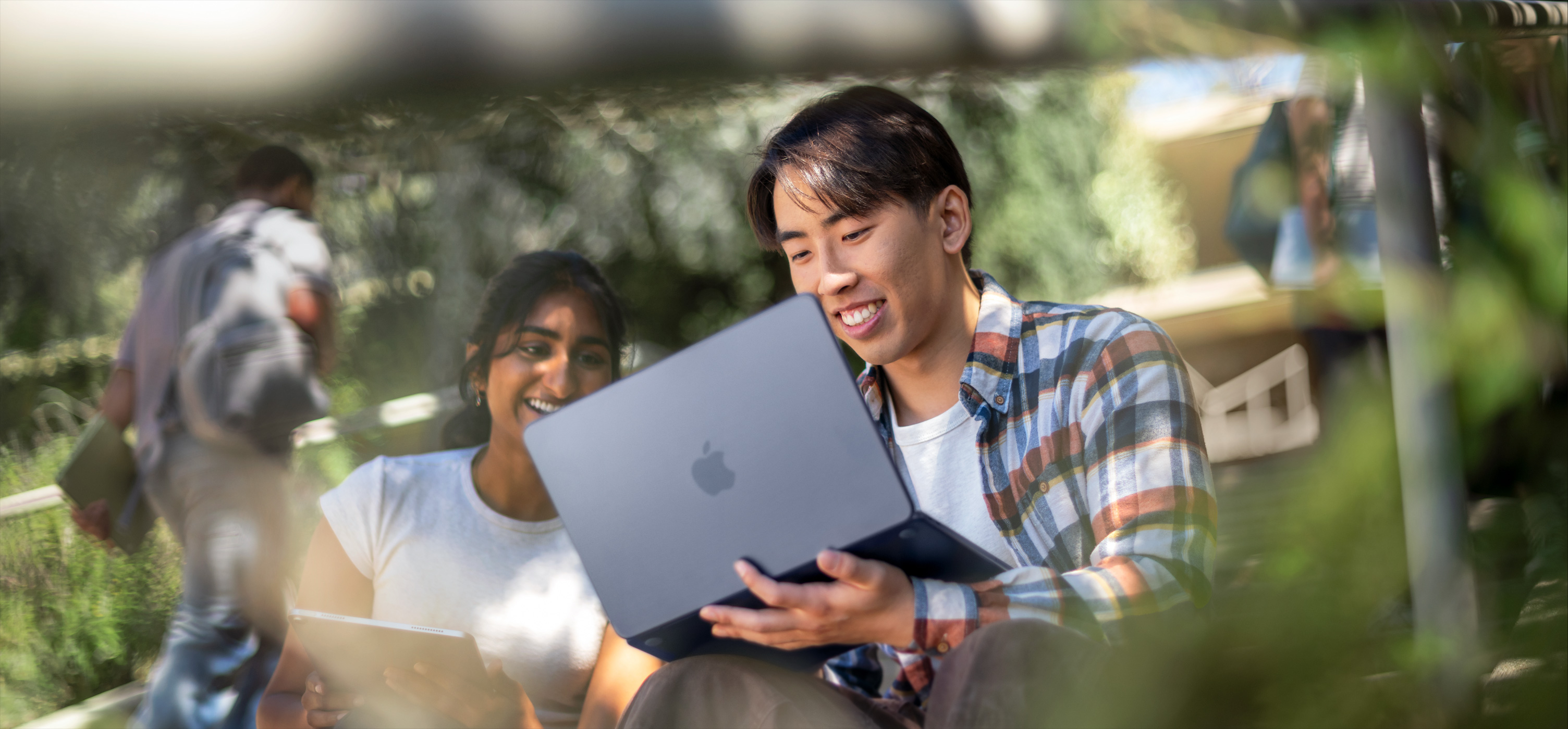 Zwei lächelnde Studierende nutzen im Freien ein MacBook, eine Person hält es auf dem Schoß, während beide gemeinsam an einer Aufgabe arbeiten