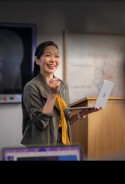 Eine Professorin hält in einem Hörsaal eine Präsentation für ihre Studierenden und hat dabei ein MacBook in Silber in der Hand, was die Performance und lange Batterielaufzeit zeigt