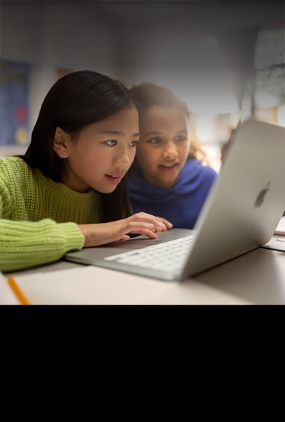 Zwei junge Schülerinnen sitzen an einem Schreibtisch in einem Klassenzimmer und arbeiten konzentriert an einem MacBook