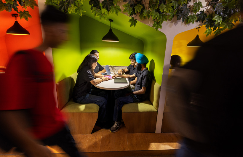 In a modern space with colourful lighting, four students at a booth work in pairs on their shared MacBook.