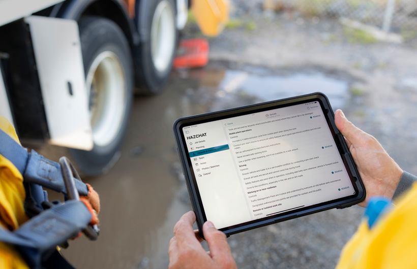 In un cantiere, accanto a una persona in piedi vicino a una pozzanghera, vengono mostrate due mani che reggono un iPad con l’app HazChat aperta; si nota il menu con le sezioni dedicate a dettagli del lavoro, pianificazione, pericoli e controlli