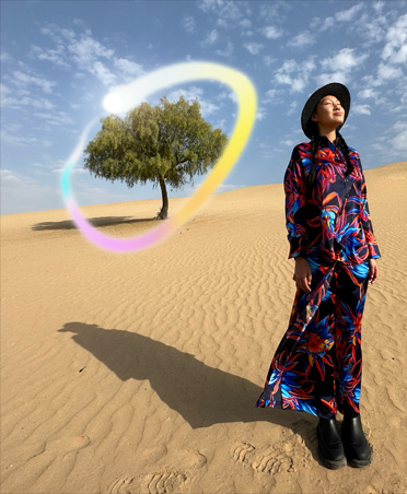 Bild einer Frau in einem mehrfarbigen Kleid, die in einer Wüstenlandschaft mit einem einsamen Baum im Hintergrund steht, der von einem leuchtenden Regenbogenverlauf umgeben ist, um die Apple Intelligence Fähigkeit zum Löschen von Hintergrundobjekten zu zeigen
