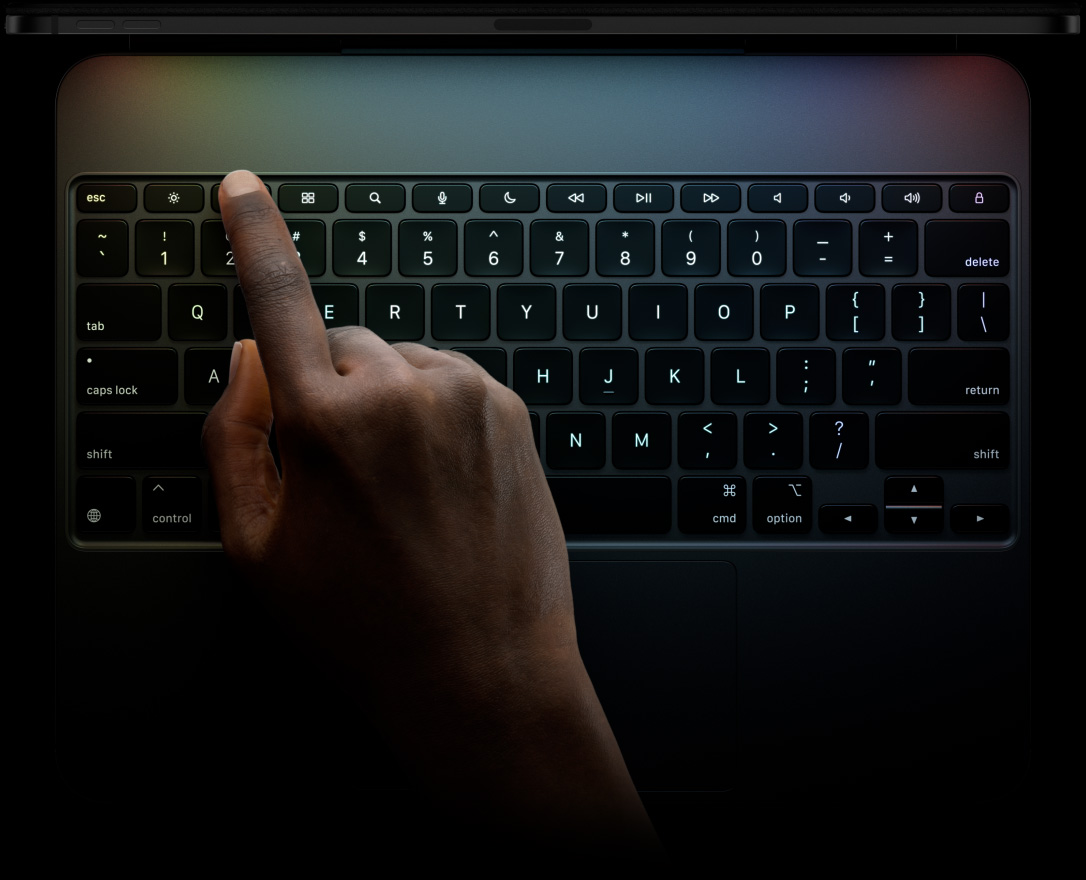 橫向放置的太空黑 iPad Pro,展示頂部邊緣輪廓,連接至精妙鍵盤;正面展示橫向放置的鍵盤,黑色背光鍵,專用功能鍵列:Escape 鍵、Tab 鍵、Caps Lock 鍵、Shift 鍵、Control 鍵、Option 鍵、Delete 鍵、Return 鍵;倒 T 形排列的方向鍵和內置觸控板。