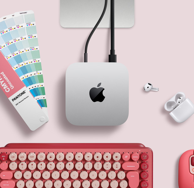 Vista desde arriba, Mac mini, aluminio color plata, esquinas redondeadas, logo de Apple negro brillante en el centro, conectada con un cable, base de un monitor Studio Display parcialmente visible, AirPods 4, muestra de una paleta de colores Pantone, teclado y mouse Logitech rojos