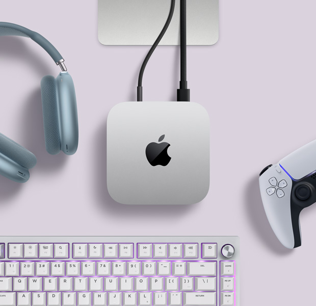 Vista desde arriba, Mac mini, aluminio color plata, esquinas redondeadas, logo de Apple negro brillante en el centro, conectada con un cable, base de un monitor Studio Display parcialmente visible, AirPods Max 2, color azul, control DualSense para PlayStation, color blanco, teclado blanco