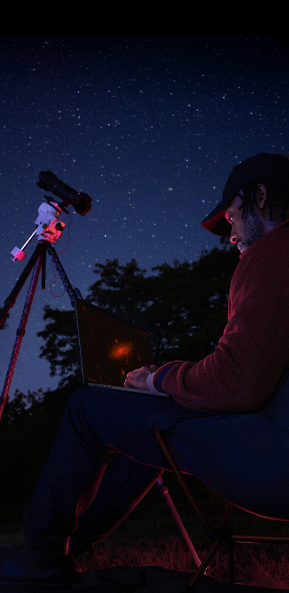 Dehors en pleine nuit, une personne tient sur ses genoux un MacBook Pro connecté à un appareil photo installé sur un trépied et pointé vers le ciel étoilé.