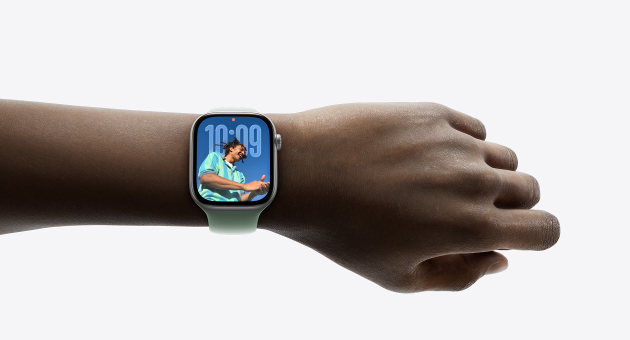 Lähikuva hopeanvärisestä Apple Watchista ja silikonikumirannekkeesta henkilön ranteessa, näkyvissä uusi Liquid Glass ‑käyttöliittymä