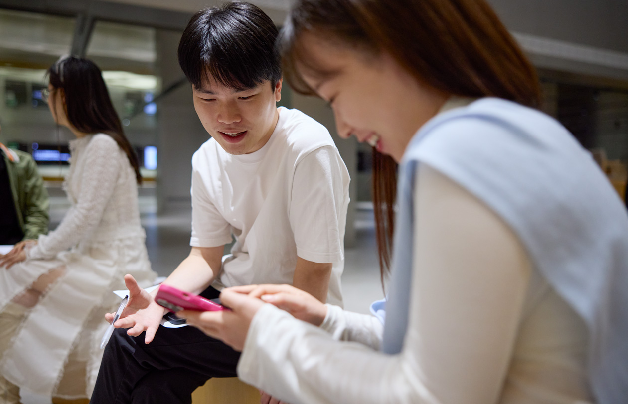2人の人物が下を向き、片方の人が手元で操作しているiPhoneを一緒に見ながら話をしている