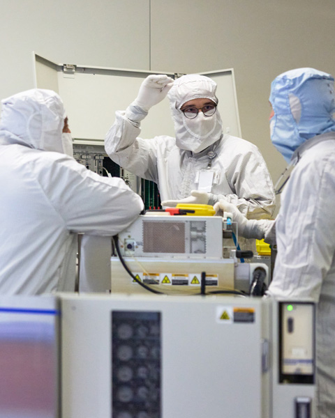 フルカバーの作業服を着た3人の従業員が、シリコン製造機の周りに立ち、一緒に作業している