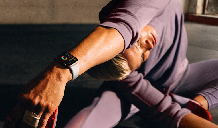 Một người phụ nữ đang ngồi trên sàn duỗi tay chạm vào chân trong khi đeo Apple Watch Series 10, màn hình hiển thị Bài Tập, Hạ Nhiệt, Hoạt Động, Vòng Hoạt Đông